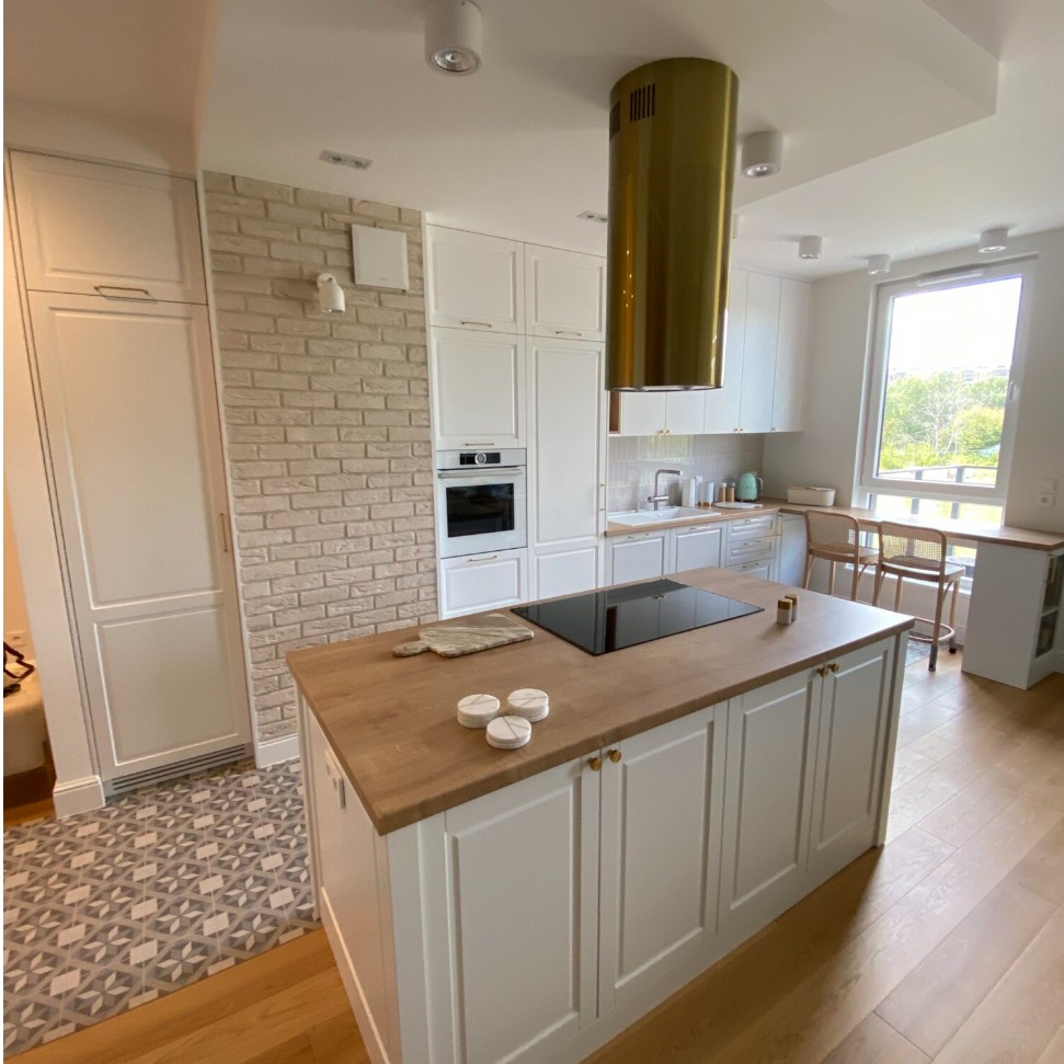 Jasna kuchnia z wyspą, drewnianym blatem i złotym okapem cylindrycznym, ściana z cegły i geometryczna podłoga w strefie wejścia.