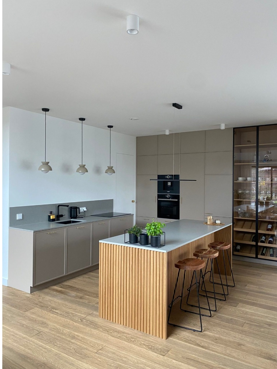 Jasna kuchnia z wyspą wykończoną pionowymi listwami, drewniane hokery i przeszklona witryna z winami w tle.