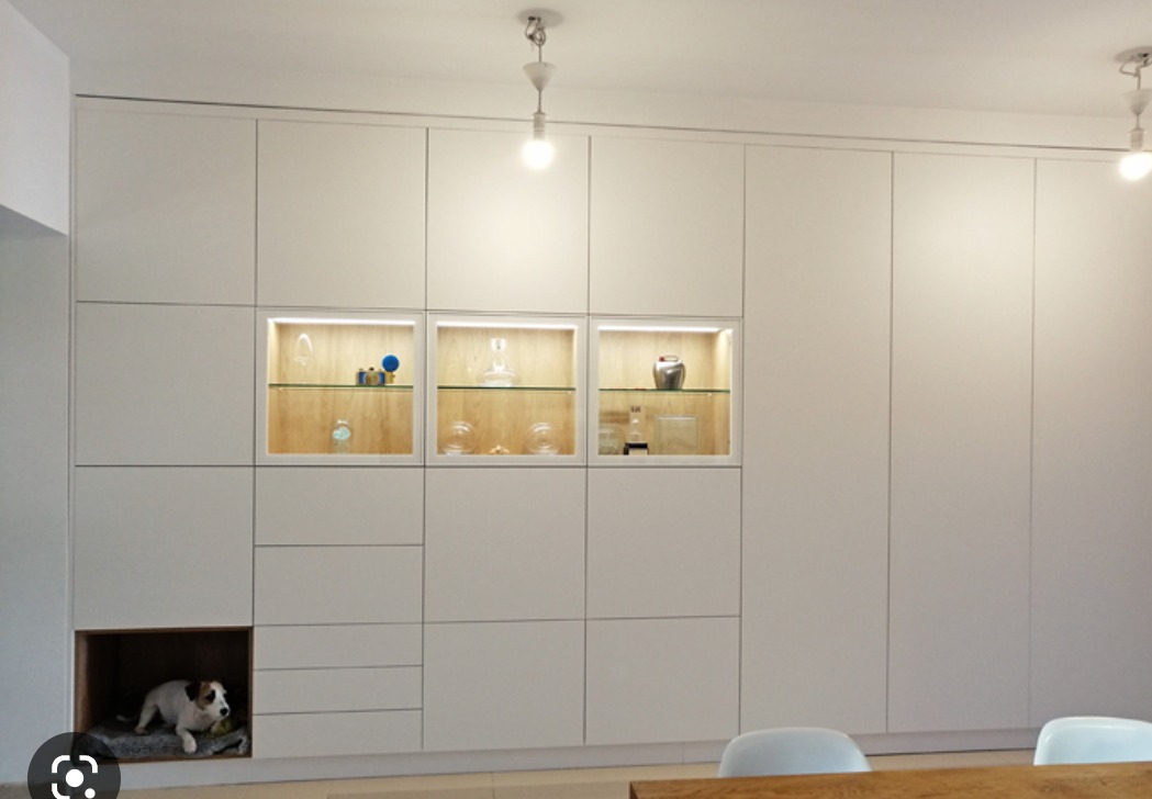 Biała zabudowa meblowa z przeszklonymi witrynami i legowiskiem dla psa w dolnej części, oświetlenie punktowe.