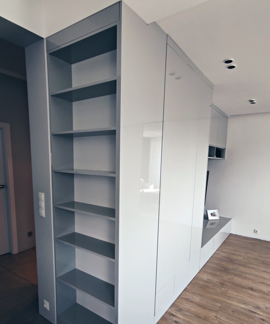 Biała zabudowa meblowa z regałem w szarym odcieniu, widoczne półki, gładkie fronty szafek, drewniana podłoga w tle.