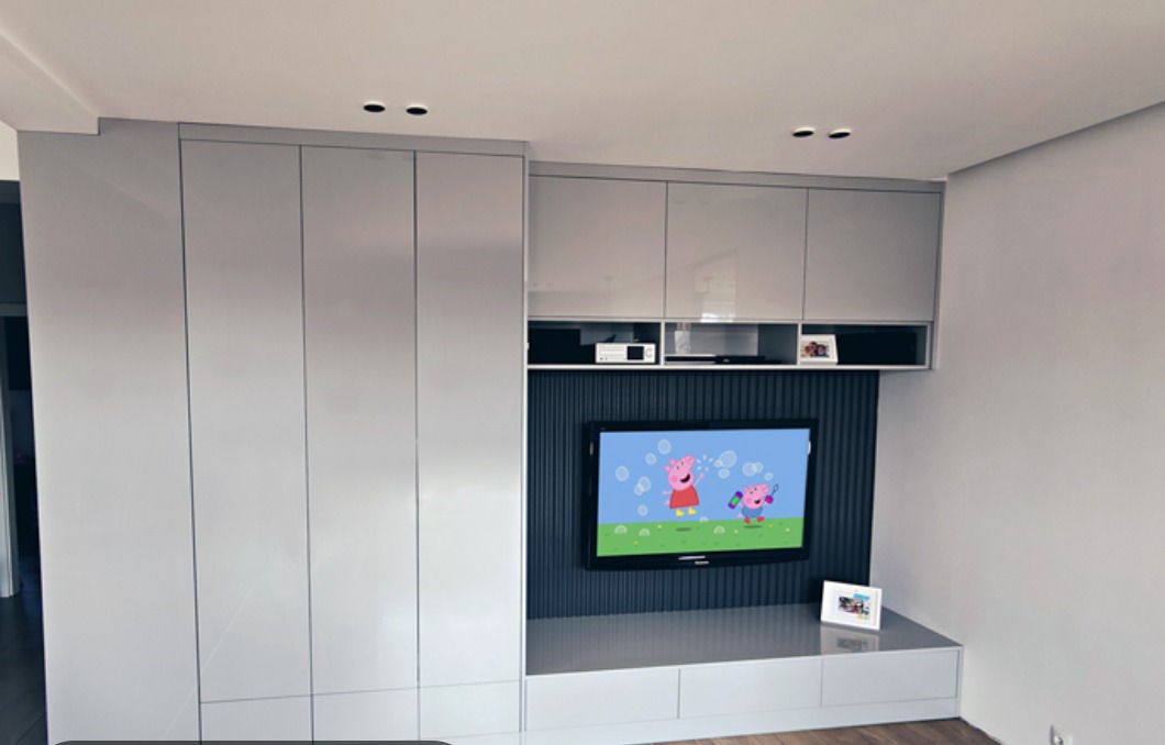 Szara zabudowa ścienna z szafkami i wnęką na telewizor z panelem dekoracyjnym. W górnej części szafki wiszące, na półkach dekoder i ramka ze zdjęciem. Na dolnej szafce stoi ramka ze zdjęciem.