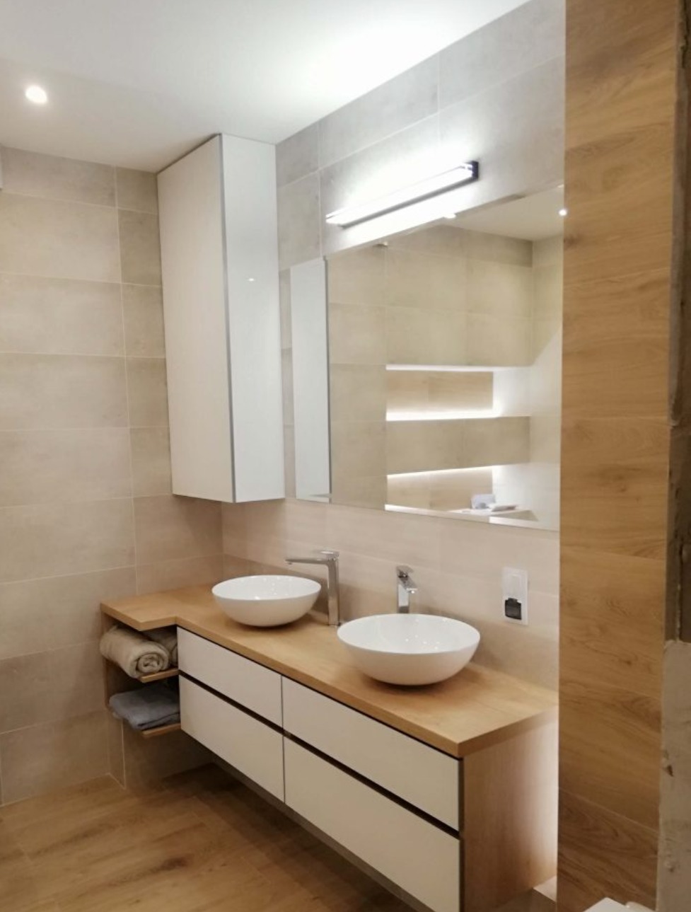 Nowoczesna łazienka z dwoma okrągłymi umywalkami nablatowymi, blatem z jasnego drewna, białymi szafkami z czarnymi uchwytami, lustrem z podświetleniem i płytkami w odcieniach beżu.