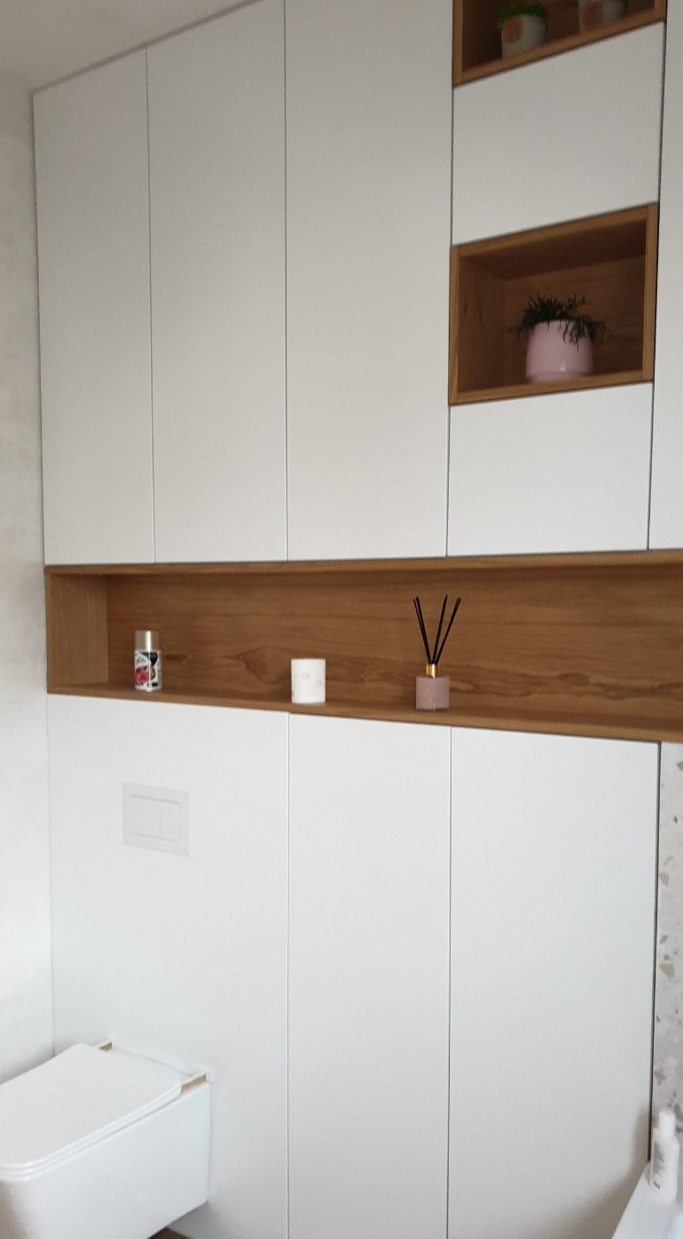Białe szafki łazienkowe na wymiar z drewnianymi wnękami, w których znajdują się dekoracje, widoczna toaleta.