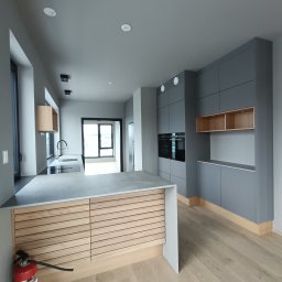 Radoslaw Szostkiewicz Montaże - Nowoczesna, minimalistyczna kuchnia z szarymi szafkami i drewnianym blatem wyspy, z widocznym czerwonym gaśnicą w rogu.