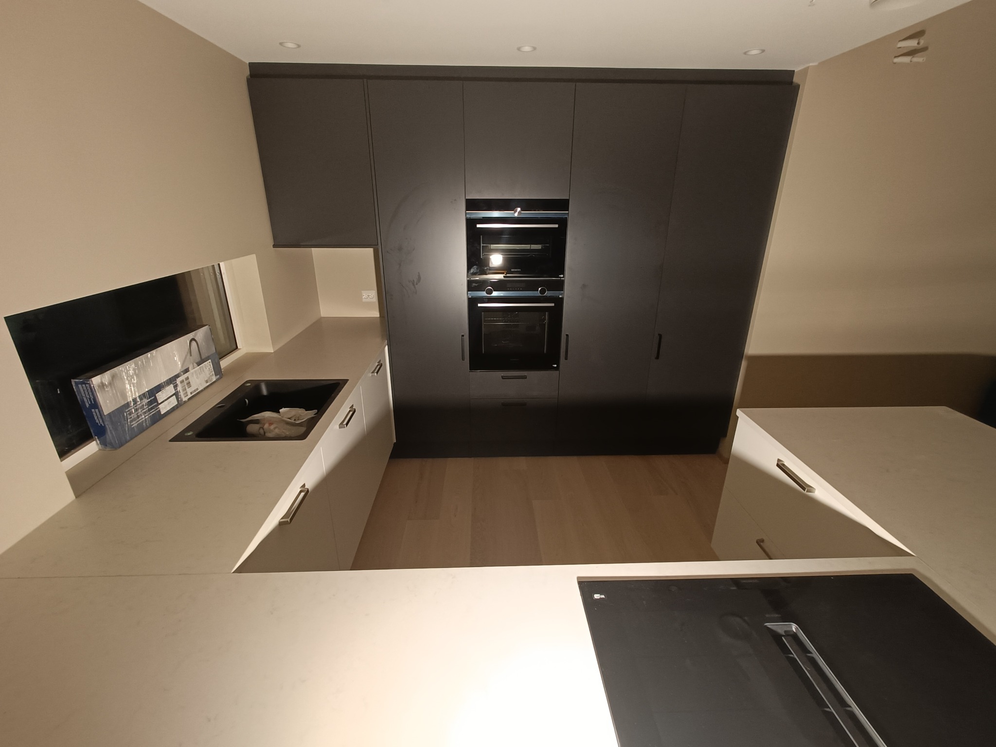 Nowoczesna kuchnia z ciemnymi, matowymi szafkami sięgającymi sufitu, zabudowanym piekarnikiem i jasnym blatem z czarnym zlewem, widok z góry na płytę indukcyjną.