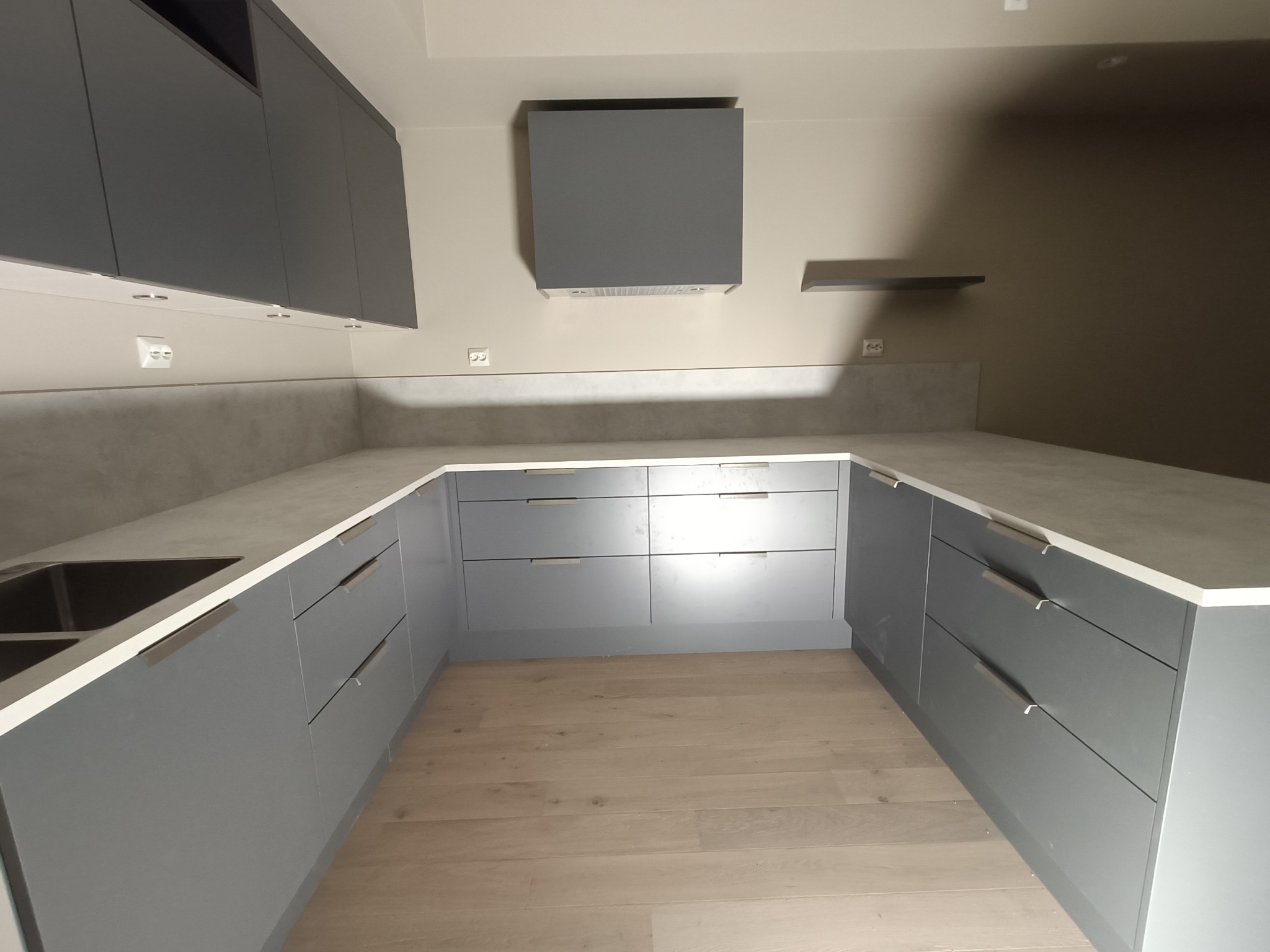 Szara kuchnia na wymiar z drewnianą podłogą; widoczny fragment zlewu, blat z szarego konglomeratu i minimalistyczny okap.