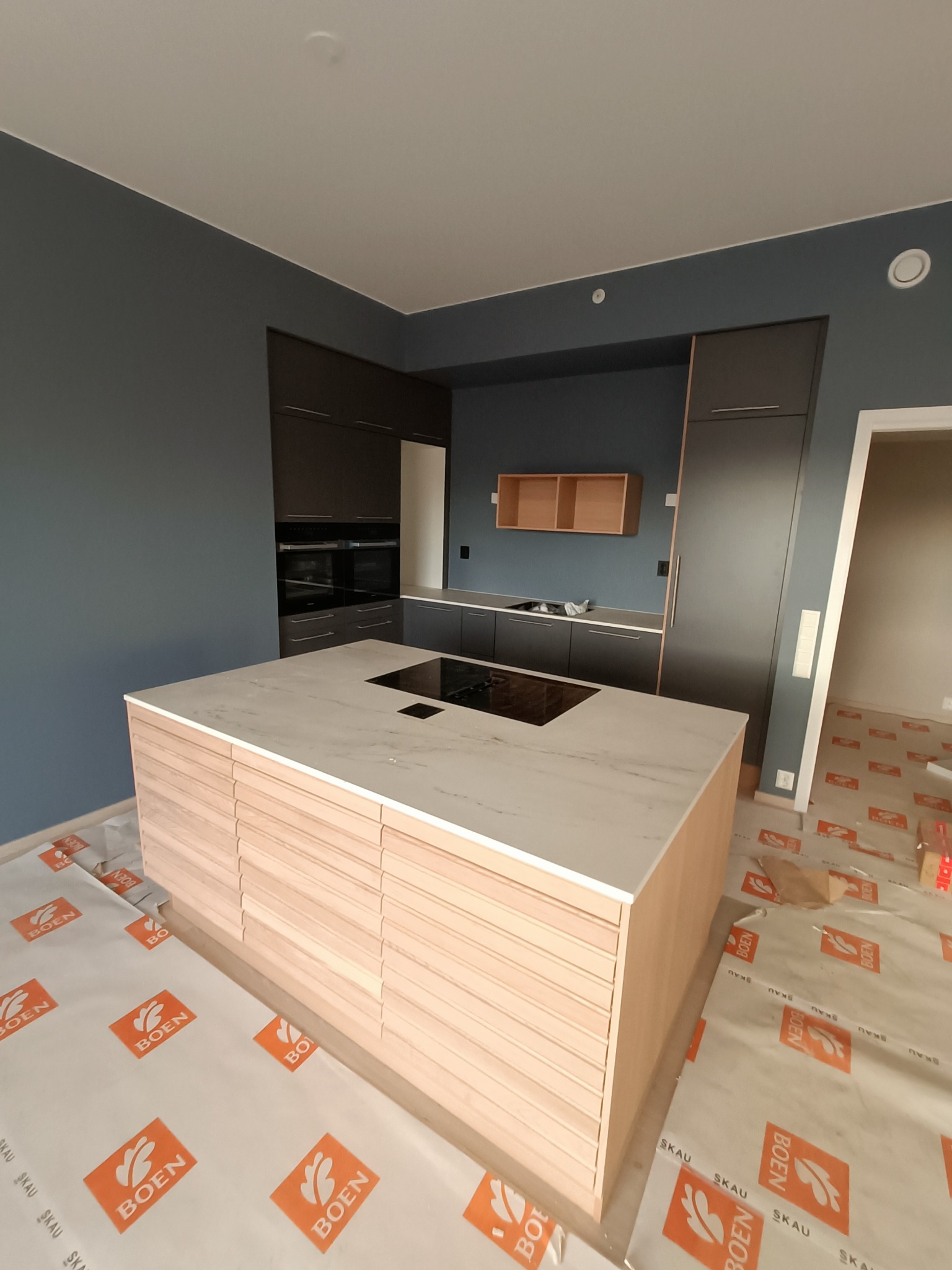 Nowoczesna kuchnia w trakcie montażu, z wyspą z drewnianymi frontami i blatem z białego marmuru, ciemnoszare szafki wiszące i stojące, podłoga zabezpieczona folią ochronną.