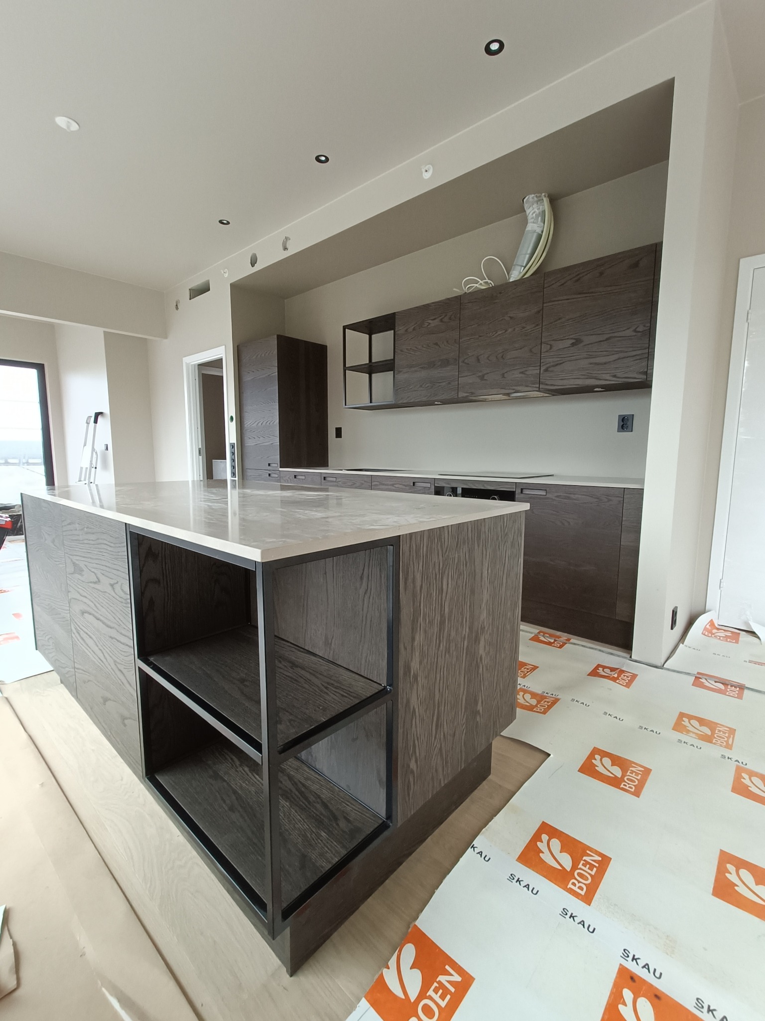 Nowoczesna kuchnia z wyspą i drewnianymi szafkami w odcieniach szarości, widoczne zabezpieczenia podłogi podczas montażu, surowy stan wykończenia.