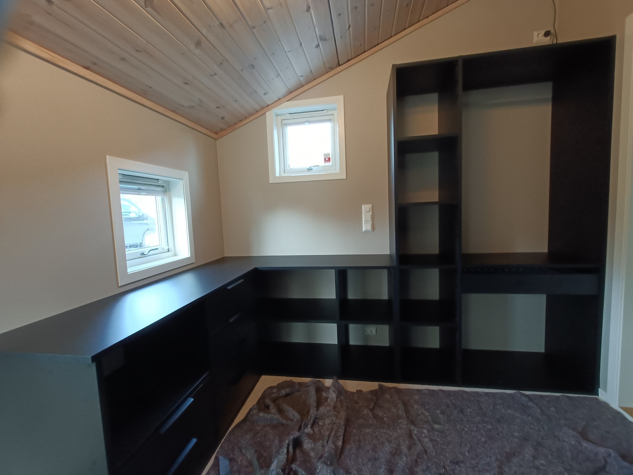 Czarna zabudowa meblowa pod skosem dachu z dwoma oknami, widoczna część szafy z półkami i drążkiem, na podłodze zabezpieczenie z folii.