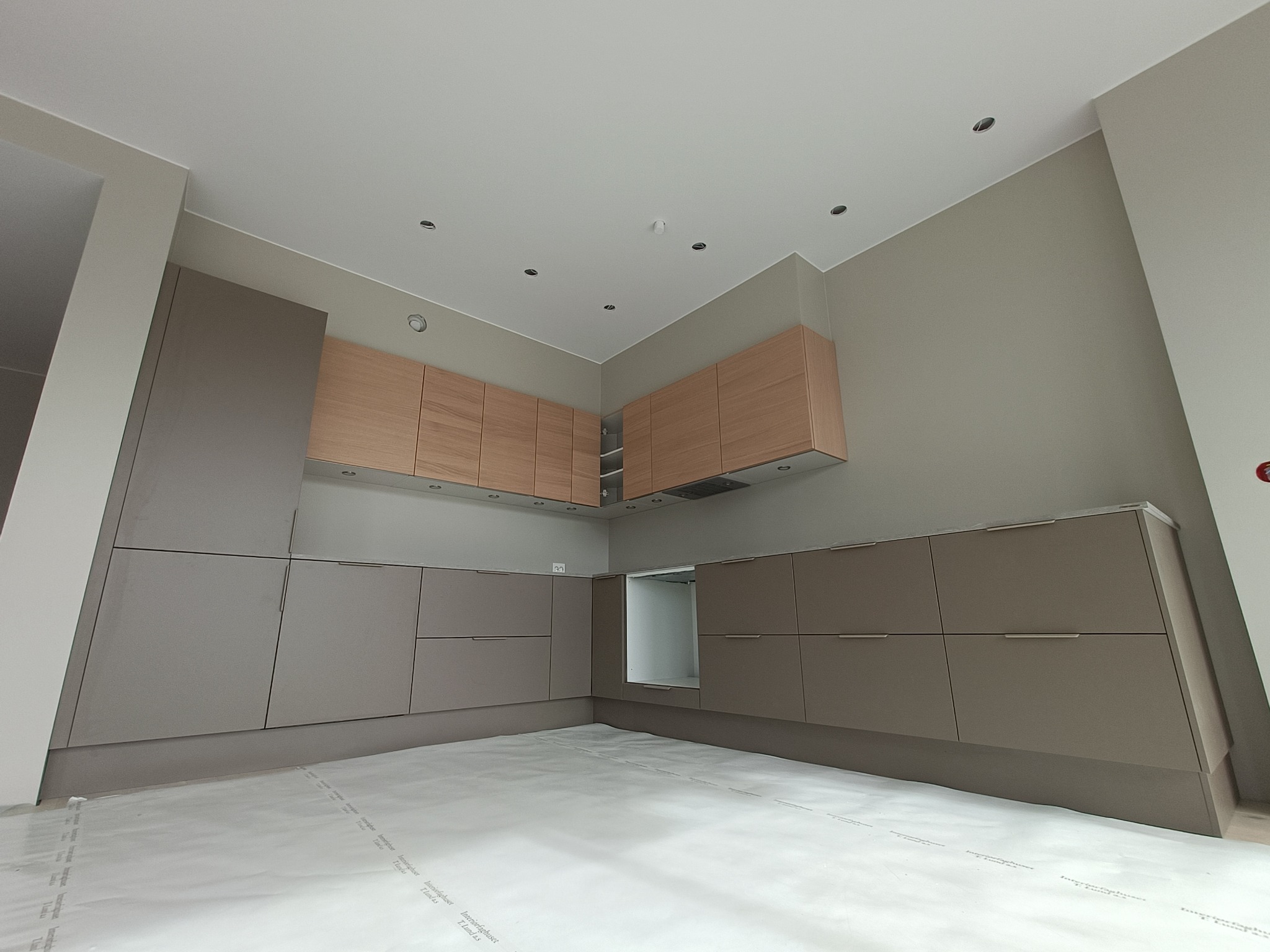 Nowoczesna kuchnia z szarymi szafkami dolnymi i drewnianymi górnymi, oświetlenie punktowe w suficie, podłoga zabezpieczona folią ochronną.