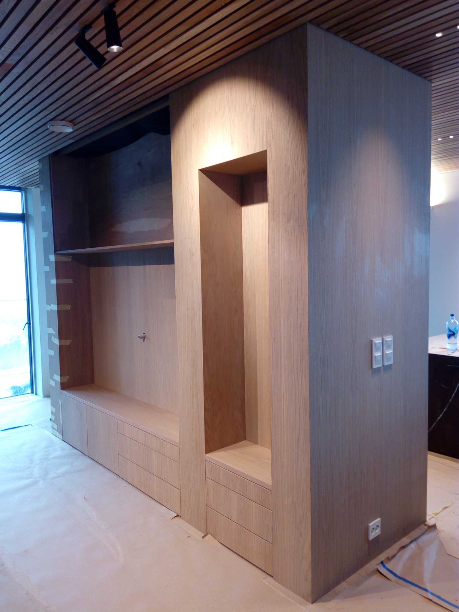 Wielofunkcyjna drewniana zabudowa w trakcie montażu: regał z półkami, szafki z szufladami i wnęka, surowe wykończenie, zabezpieczona podłoga.