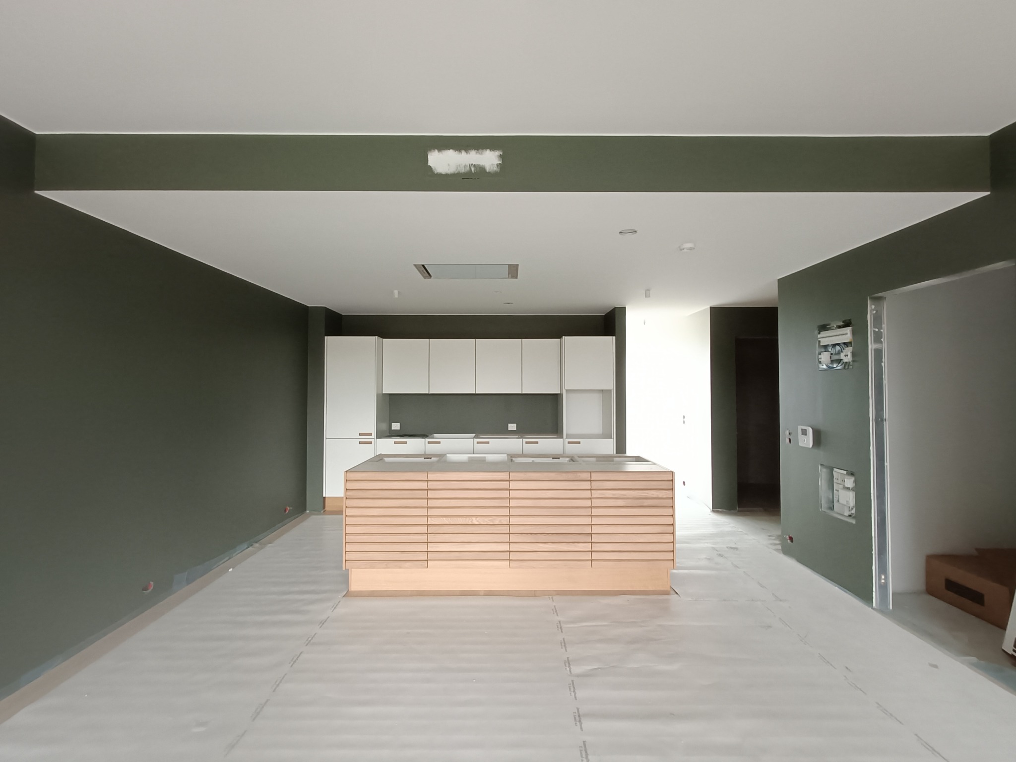 Wnętrze w trakcie wykańczania: widok na białe szafki kuchenne i drewnianą wyspę z poziomymi listwami, podłoga zabezpieczona folią, ściany w kolorze oliwkowym, widoczne puszki elektryczne.