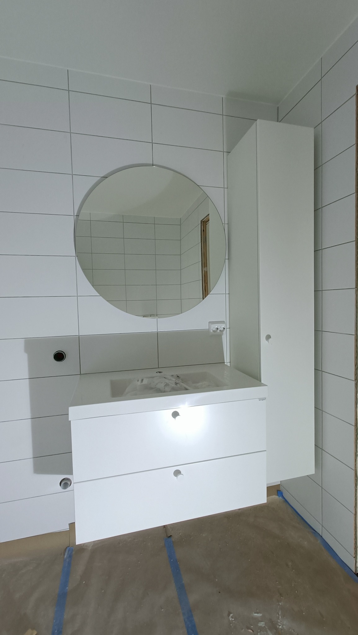 Biała szafka łazienkowa z dwoma szufladami i okrągłym lustrem, zamontowana na tle białych płytek, z widocznymi instalacjami pod umywalkę i wysoką szafką po prawej stronie. Podłoga zabezpieczona...