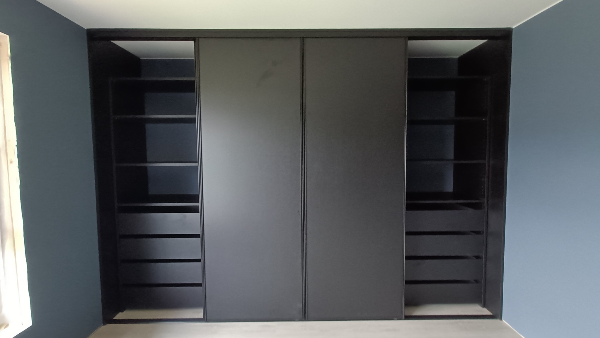 Czarna szafa wnękowa z przesuwanymi drzwiami, z widocznymi półkami i szufladami po bokach, na tle ściany w odcieniach szarości i błękitu.