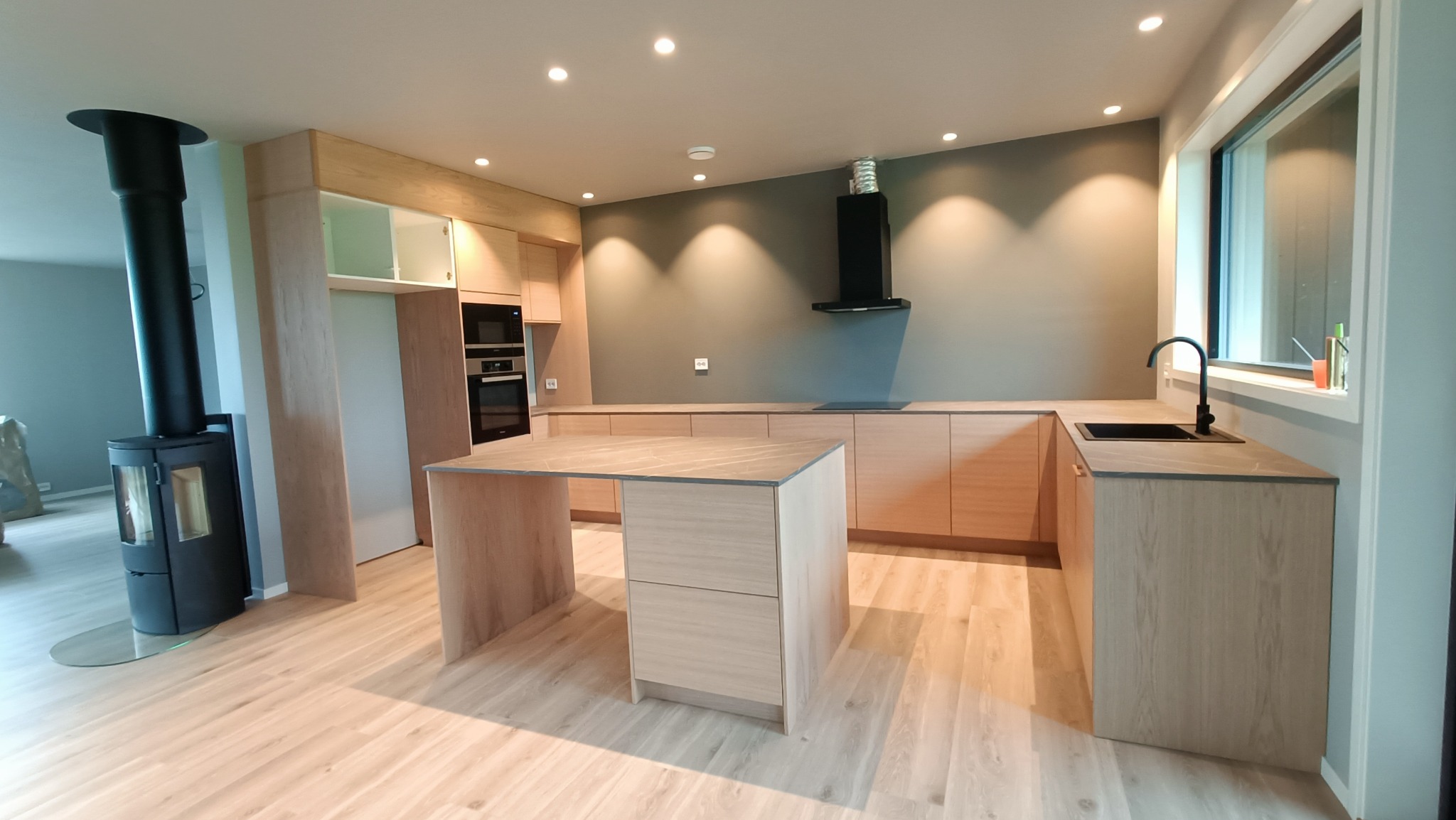 Jasna kuchnia na wymiar z drewnianymi frontami, wyspą kuchenną z blatem imitującym marmur, czarną armaturą i piecem wolnostojącym.