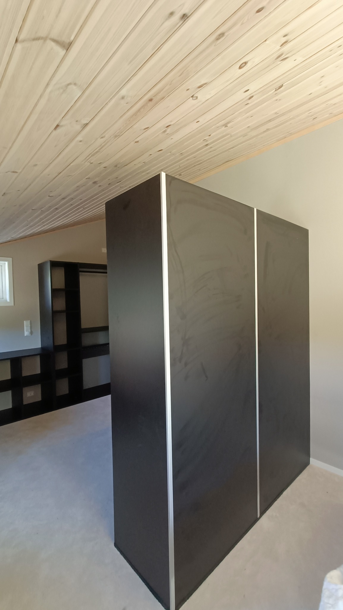 Czarna, minimalistyczna garderoba z przesuwnymi drzwiami w pomieszczeniu z drewnianym sufitem i widoczną w tle otwartą zabudową garderoby.