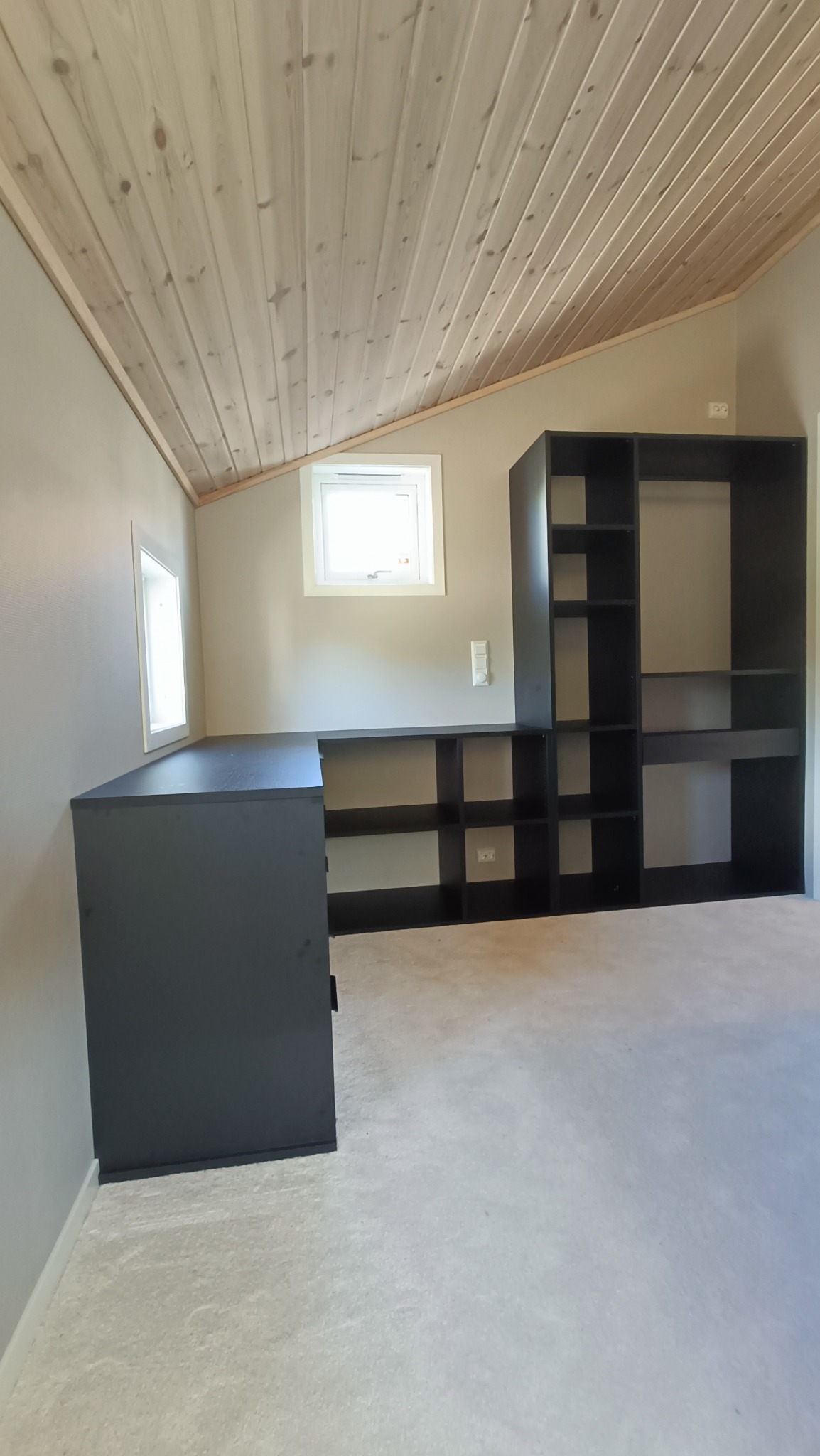 Czarna zabudowa meblowa z biurkiem i regałami pod skosnym, drewnianym sufitem na poddaszu, widoczne gniazdka elektryczne na ścianie, dwa okna w białych ramach.