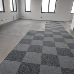 Garant-BUD - Wnętrze w stanie deweloperskim z częściowo ułożoną szarą, kwadratową wykładziną dywanową w wzór szachownicy, widoczne okna i przygotowane punkty elektryczne.