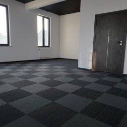 Garant-BUD - Wnętrze biurowe z szarą wykładziną dywanową w kratę, białe ściany, ciemne drzwi z pionowym srebrnym paskiem i oknami w czarnych ramach.