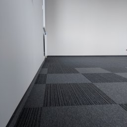 Garant-BUD - Szara podłoga wyłożona kwadratowymi płytkami dywanowymi o zróżnicowanej fakturze, widok na fragment ściany i drzwi w tle.