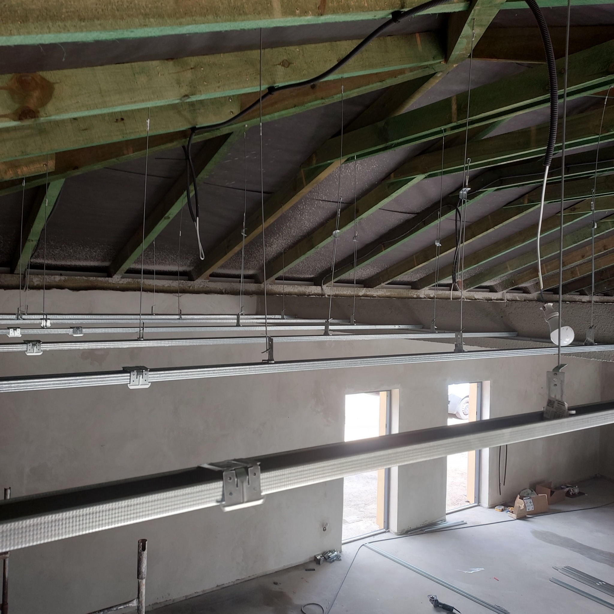 Montaż sufitu podwieszanego z widocznymi profilami CD i UD, zawieszonymi na drutach mocowanych do drewnianej konstrukcji dachu, ściany w stanie surowym.