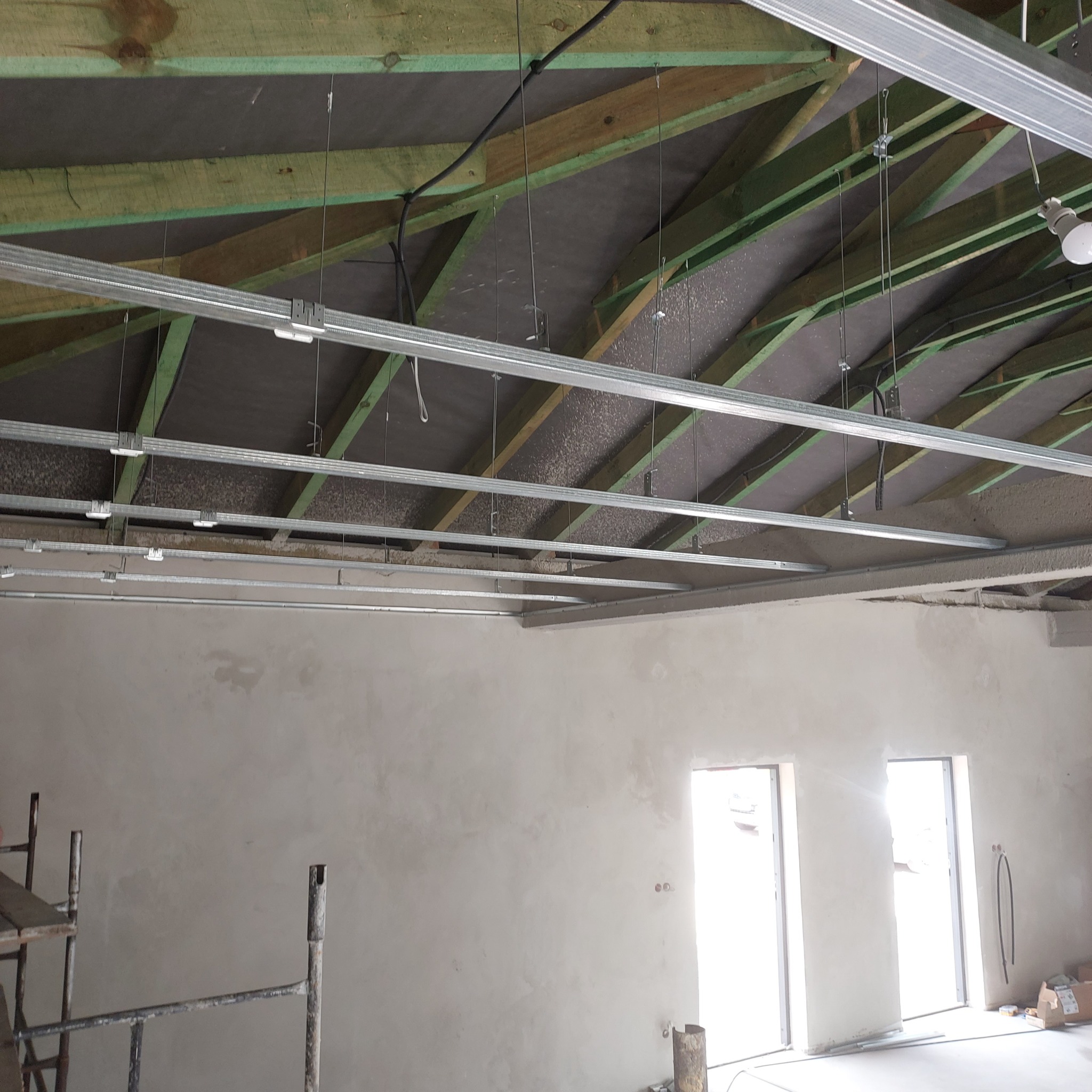 Konstrukcja sufitu podwieszanego z widocznymi profilami metalowymi, zawiesiami i elementami izolacji na drewnianej więźbie dachowej, ściany w stanie surowym, widoczne otwory okienne i drzwiowe.