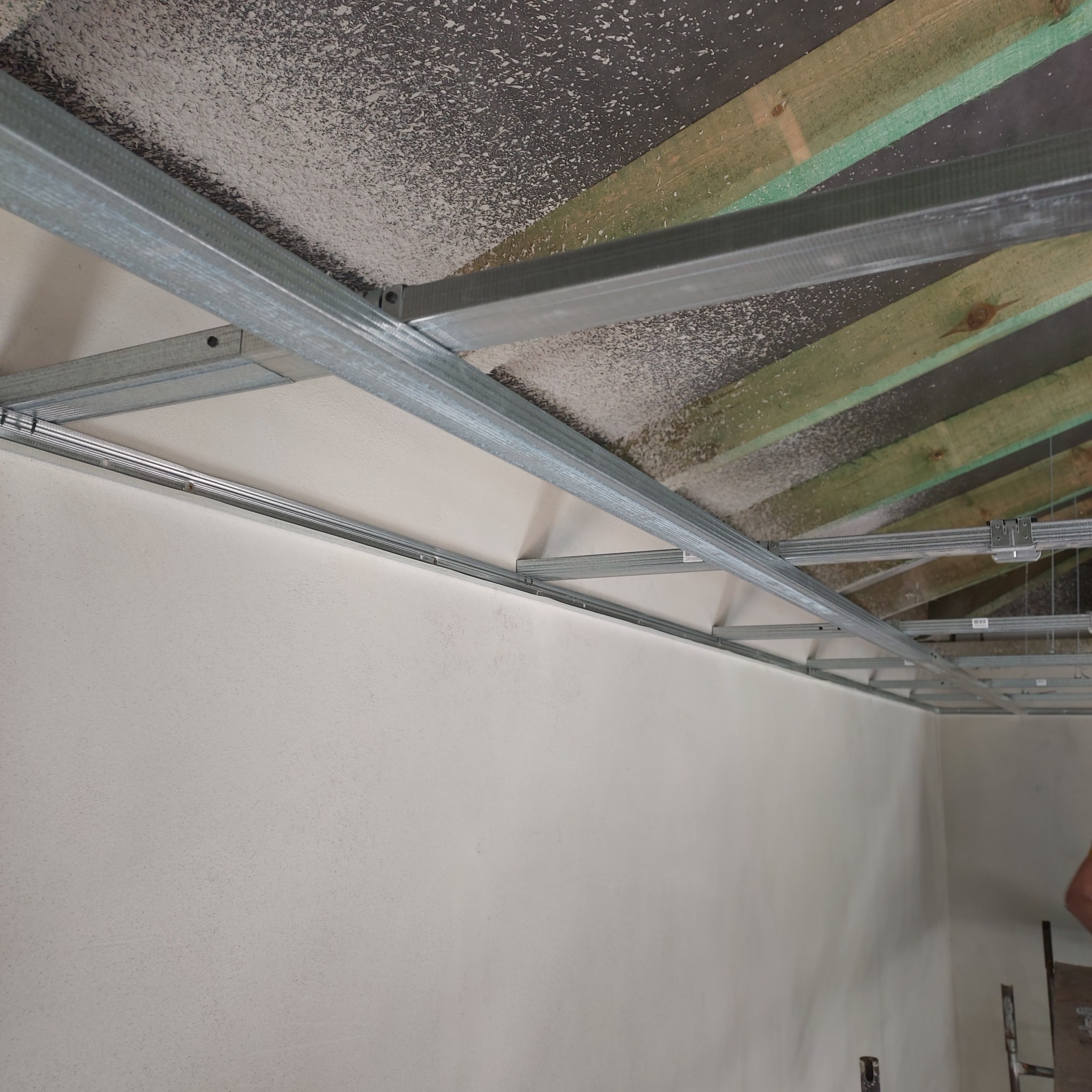 Stelaż sufitu podwieszanego z profili metalowych, widoczne drewniane elementy konstrukcyjne stropu i fragment ściany z gładzią.