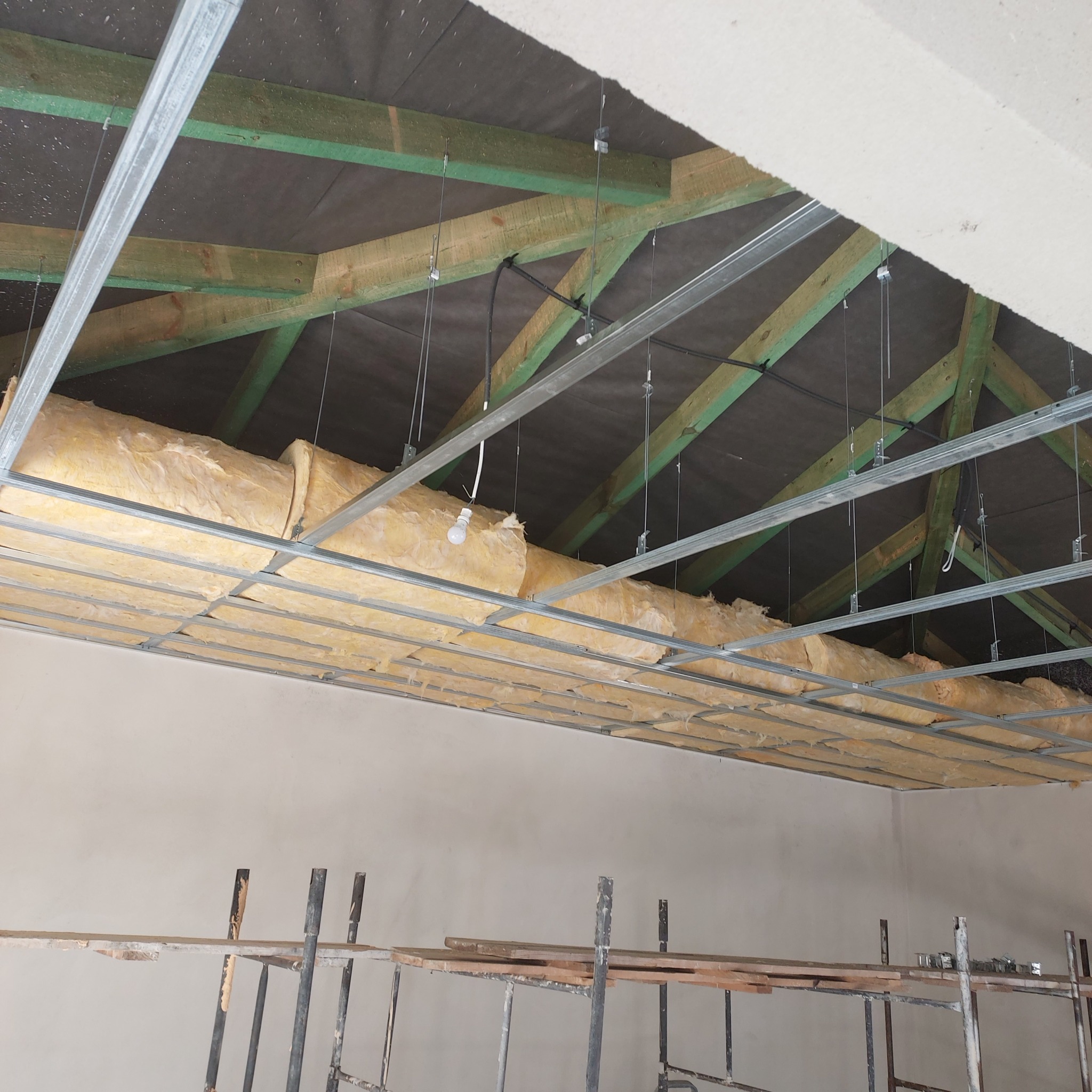 Montaż sufitu podwieszanego z izolacją z wełny mineralnej, widoczne drewniane krokwie, metalowa konstrukcja i tymczasowe oświetlenie.