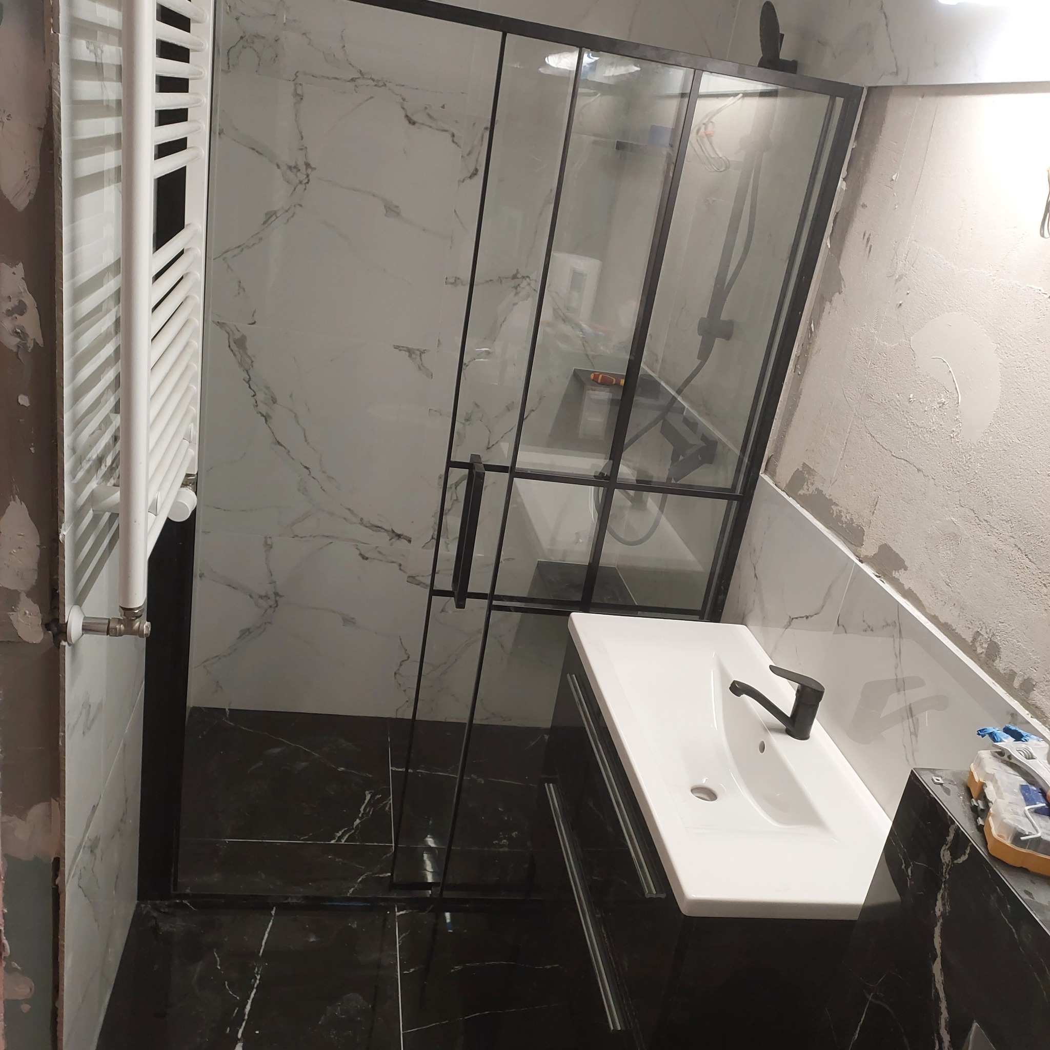 Łazienka w trakcie wykańczania: kabina prysznicowa z czarnymi profilami, biała umywalka z czarną baterią, grzejnik drabinkowy, marmurowe płytki na ścianach i podłodze, surowy fragment ściany...
