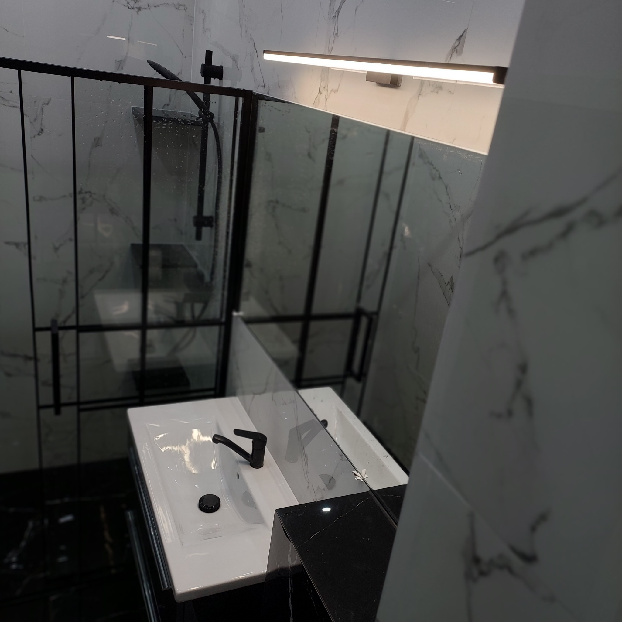Nowoczesna łazienka z czarną armaturą, kabiną prysznicową typu walk-in i marmurowymi płytkami na ścianach, oświetlona listwą LED nad lustrem.