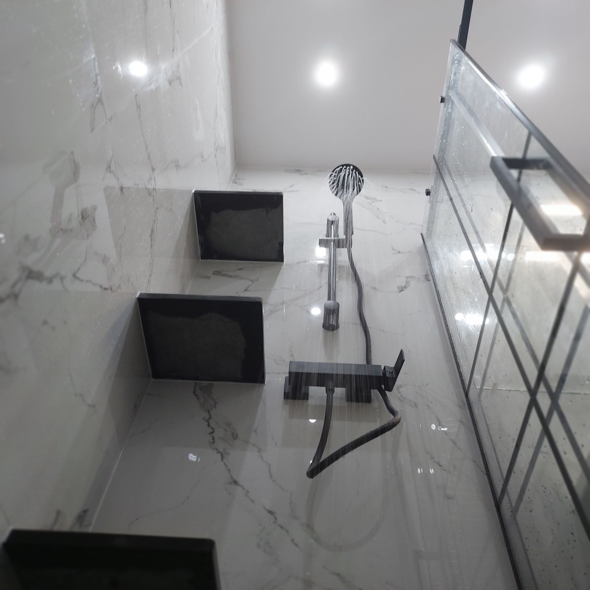 Nowoczesna kabina prysznicowa z czarną armaturą i marmurowymi płytkami, woda leje się z deszczownicy, widoczne półki wnękowe.