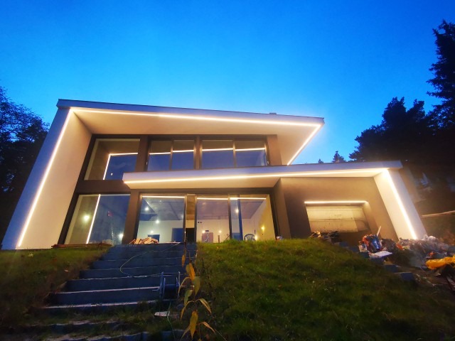 Nowoczesny dom jednorodzinny z elewacją w odcieniach szarości, oświetlony ledowymi listwami na krawędziach dachu i ścian, widok z dołu na tle wieczornego nieba.