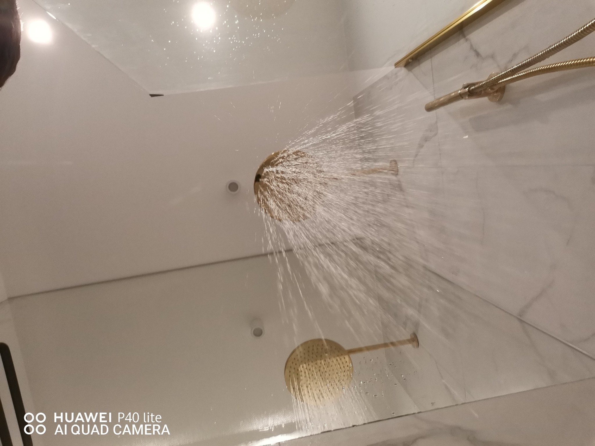 Złota deszczownica w kabinie prysznicowej w trakcie testu, woda leci z dysz pod ciśnieniem, widoczne białe ściany z marmurowym wzorem.
