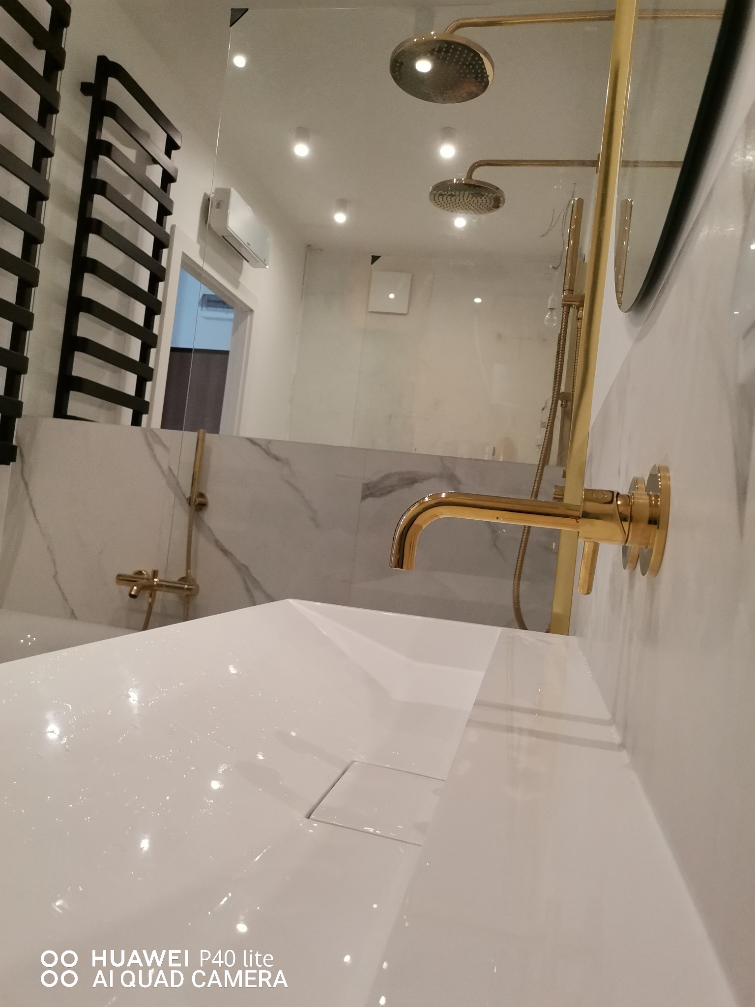 Nowoczesna łazienka z białą umywalką, złotą armaturą i czarnym grzejnikiem drabinkowym na tle marmurowej ściany.