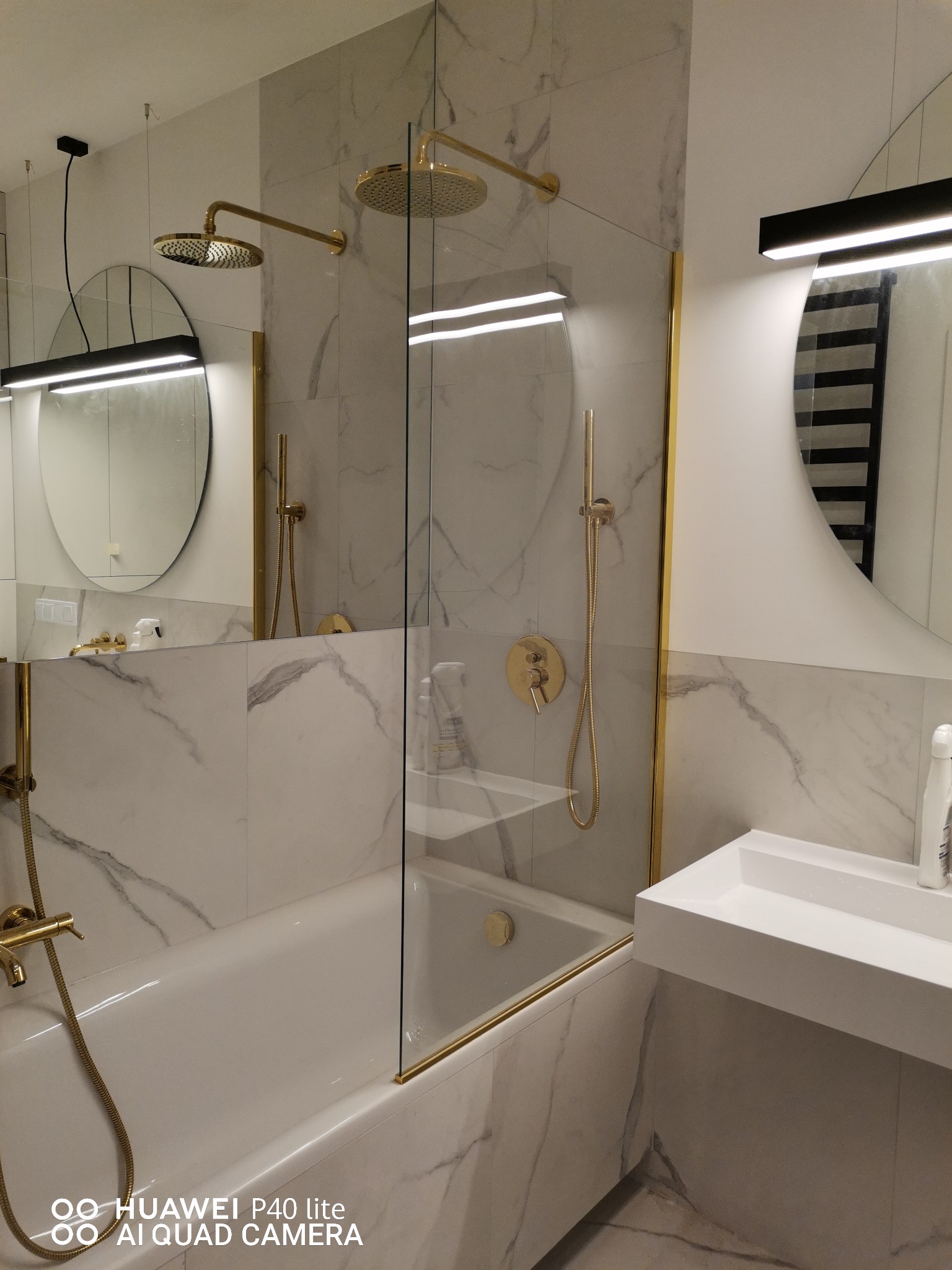 Nowoczesna łazienka z wanną i kabiną prysznicową oddzieloną szybą z złotymi ramami, marmurowe płytki na ścianach, okrągłe lustra z podświetleniem LED, złota armatura.