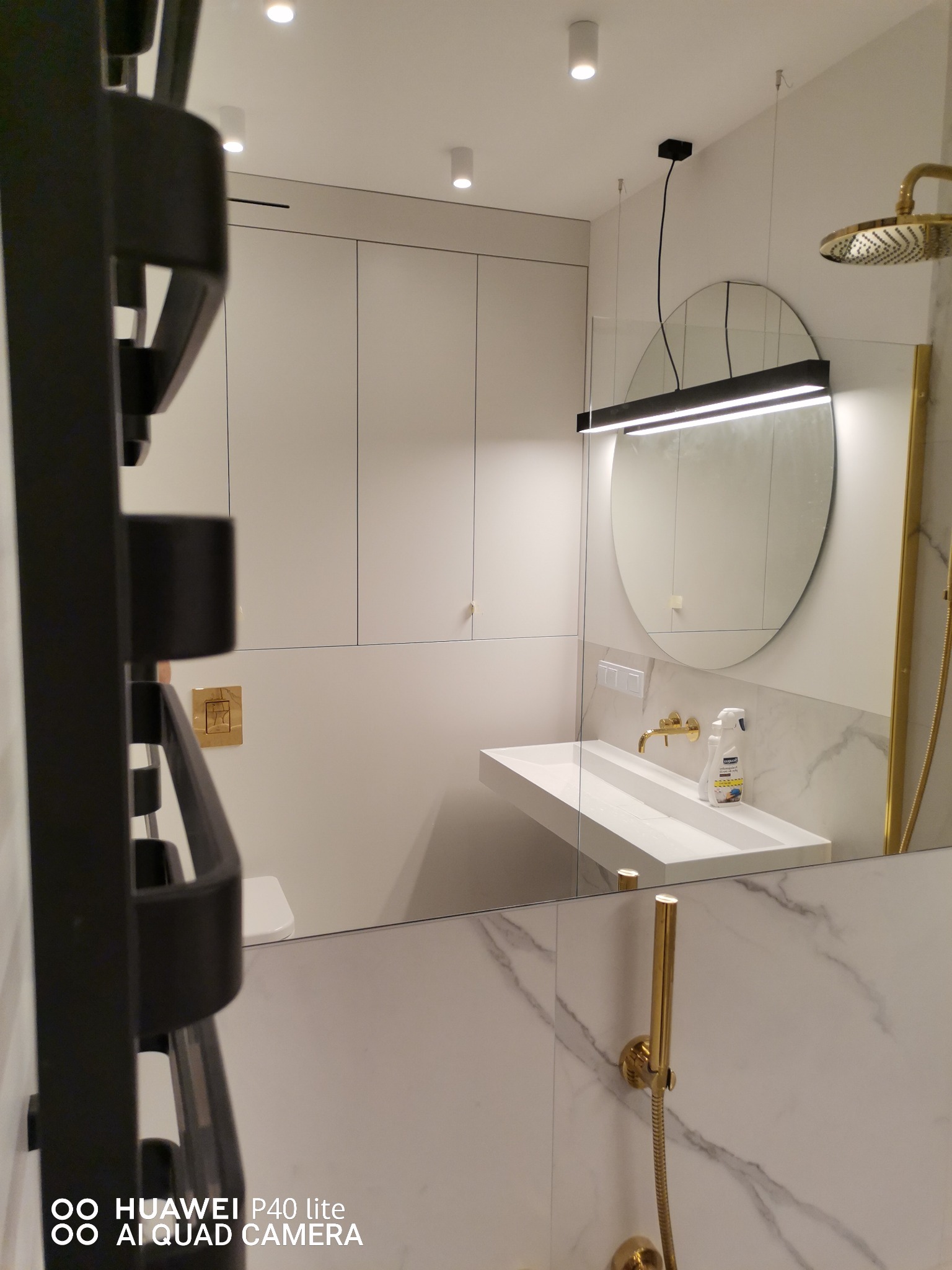 Nowoczesna łazienka z białymi szafkami, podwieszaną umywalką, okrągłym lustrem z podświetleniem LED i złotą armaturą, widziana w odbiciu lustra.