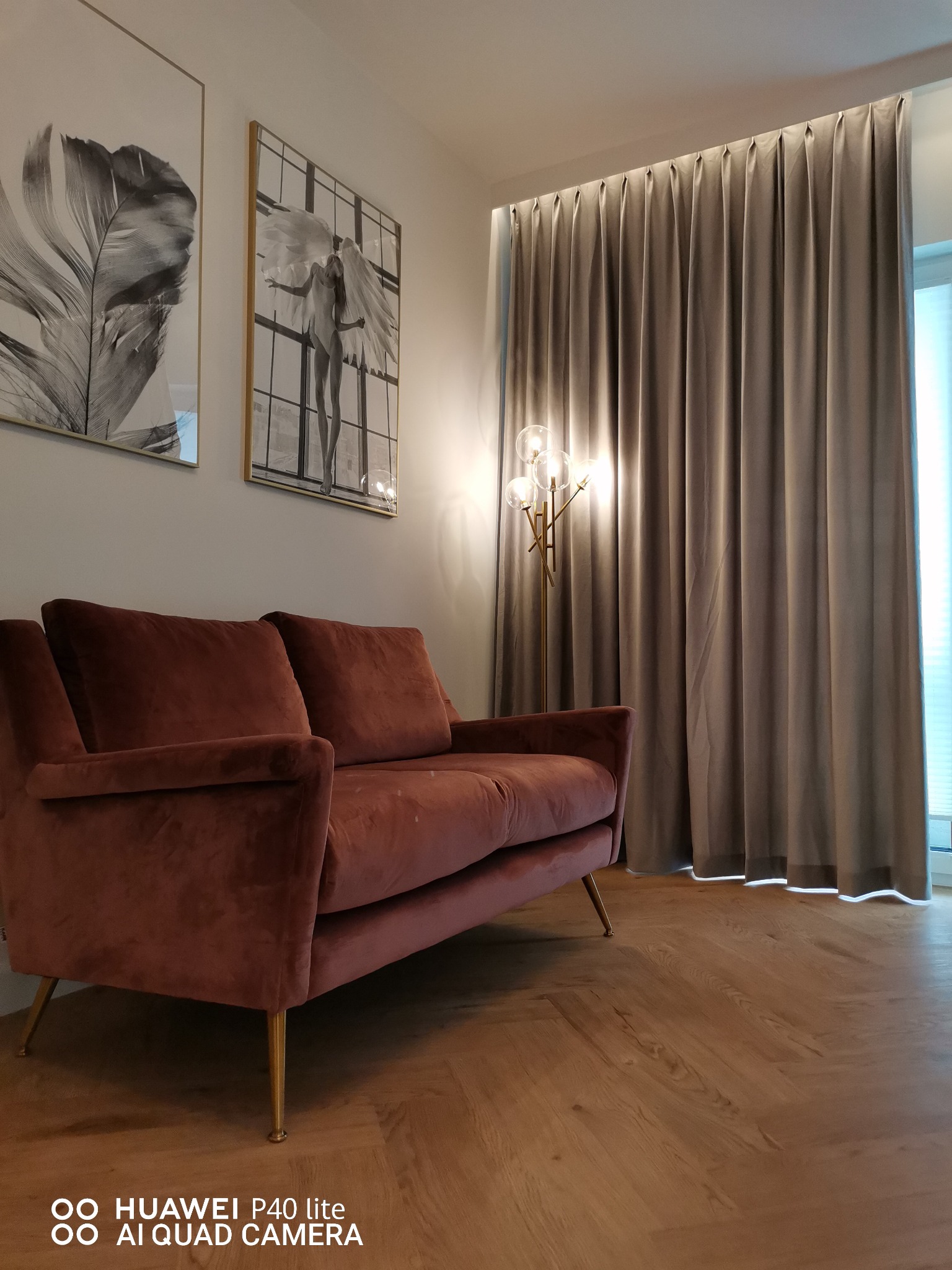 Stylowe wnętrze: bordowa sofa na złotych nóżkach, drewniana podłoga w jodełkę, szare zasłony z subtelnym oświetleniem i dekoracyjne obrazy na ścianie.