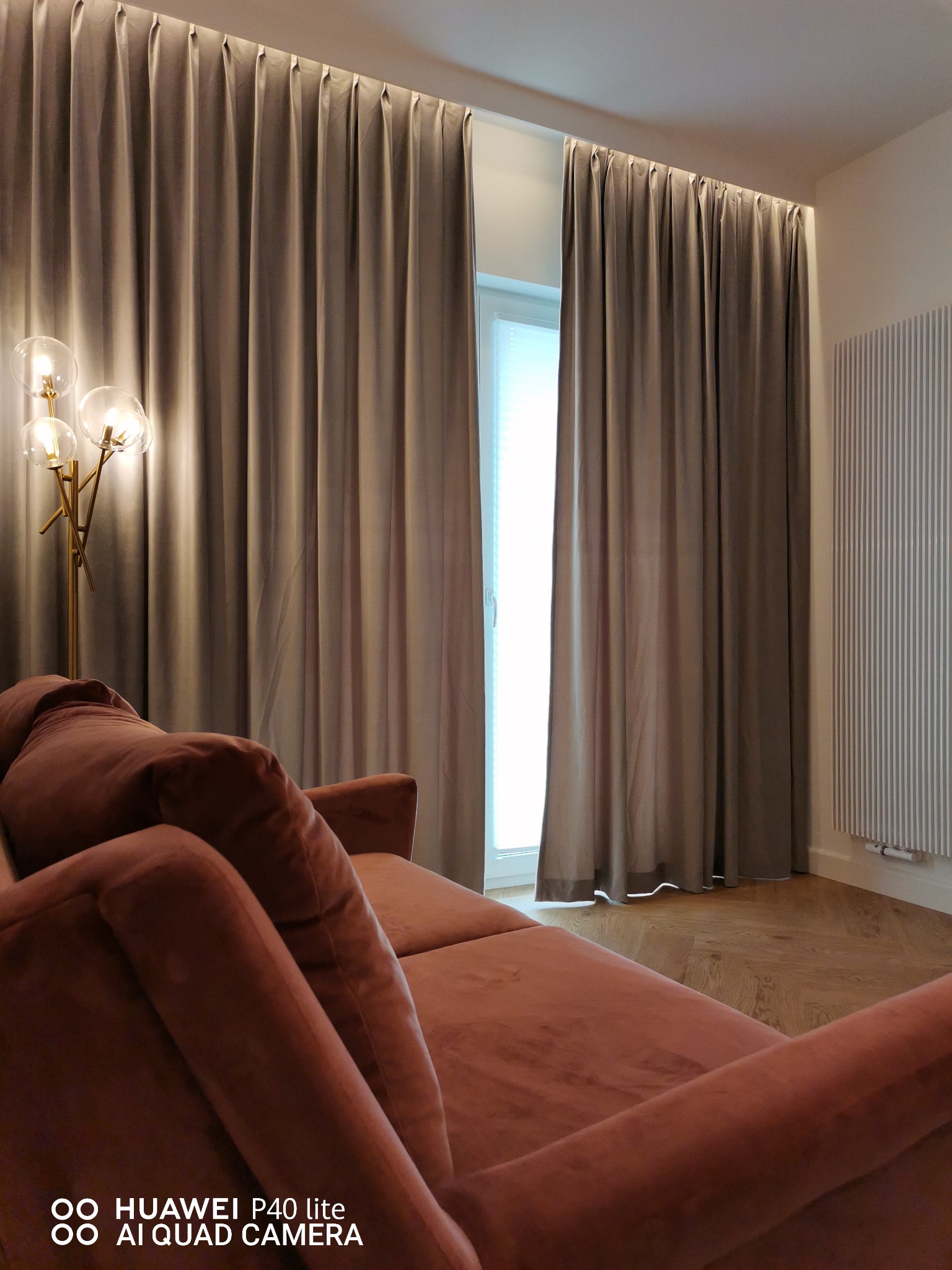 Beżowe zasłony z ukrytym karniszem sufitowym, widoczne z perspektywy pluszowej, brzoskwiniowej sofy i lampy podłogowej z trzema szklanymi kulami.