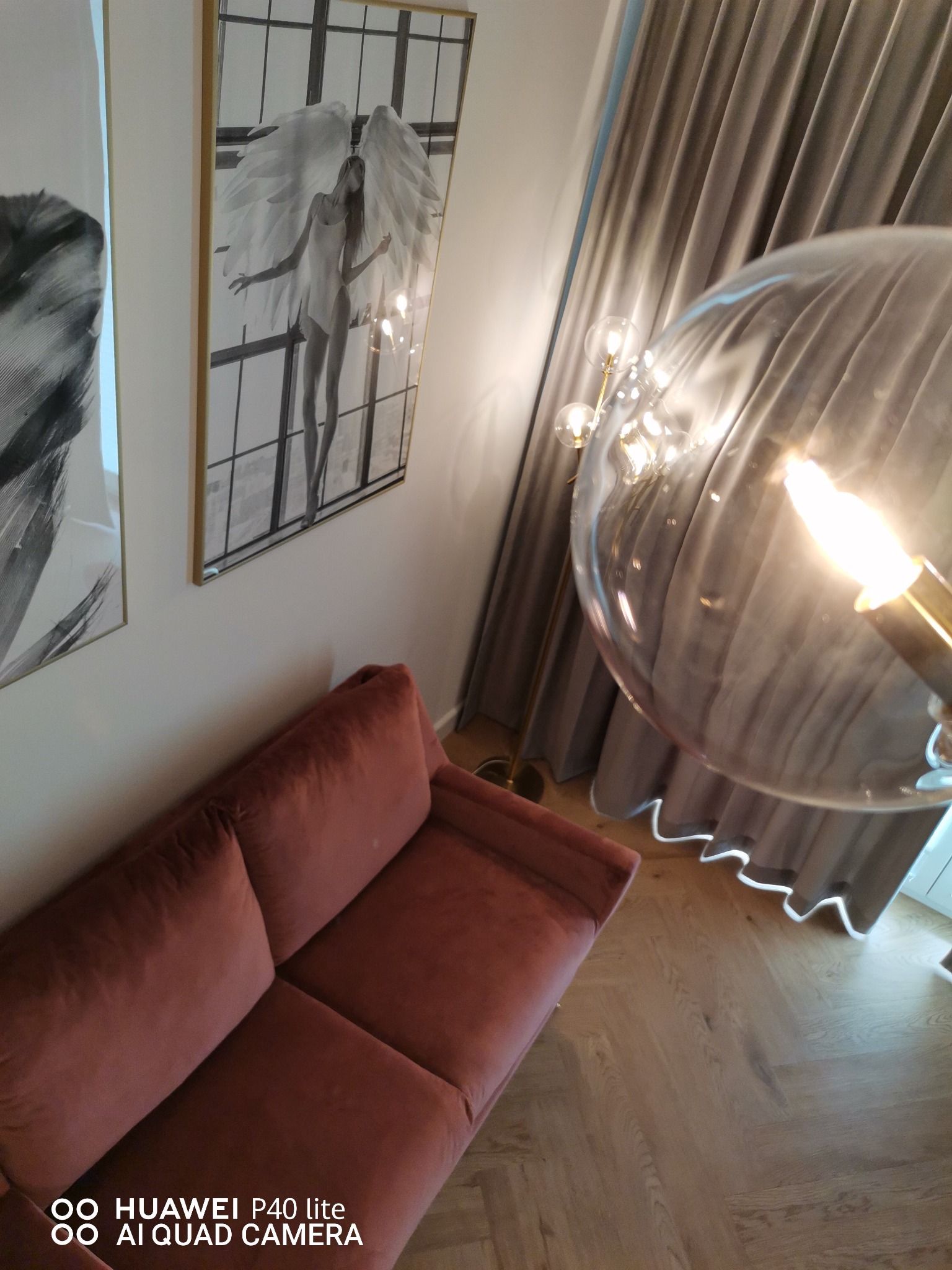 Wnętrze pokoju z aksamitną sofą w odcieniach różu, nowoczesną lampą z kulistym kloszem i grafikami na ścianie, widoczne detale podłogi z jasnego drewna.