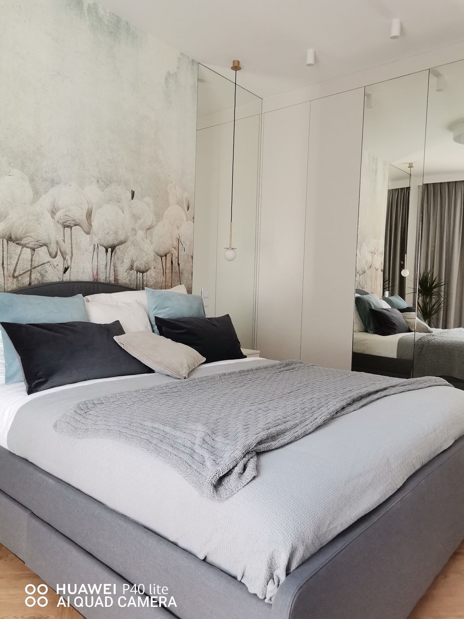 Nowoczesna sypialnia z łóżkiem w odcieniach szarości, dekoracyjną tapetą z motywem flamingów i lustrzanymi drzwiami szafy wnękowej.