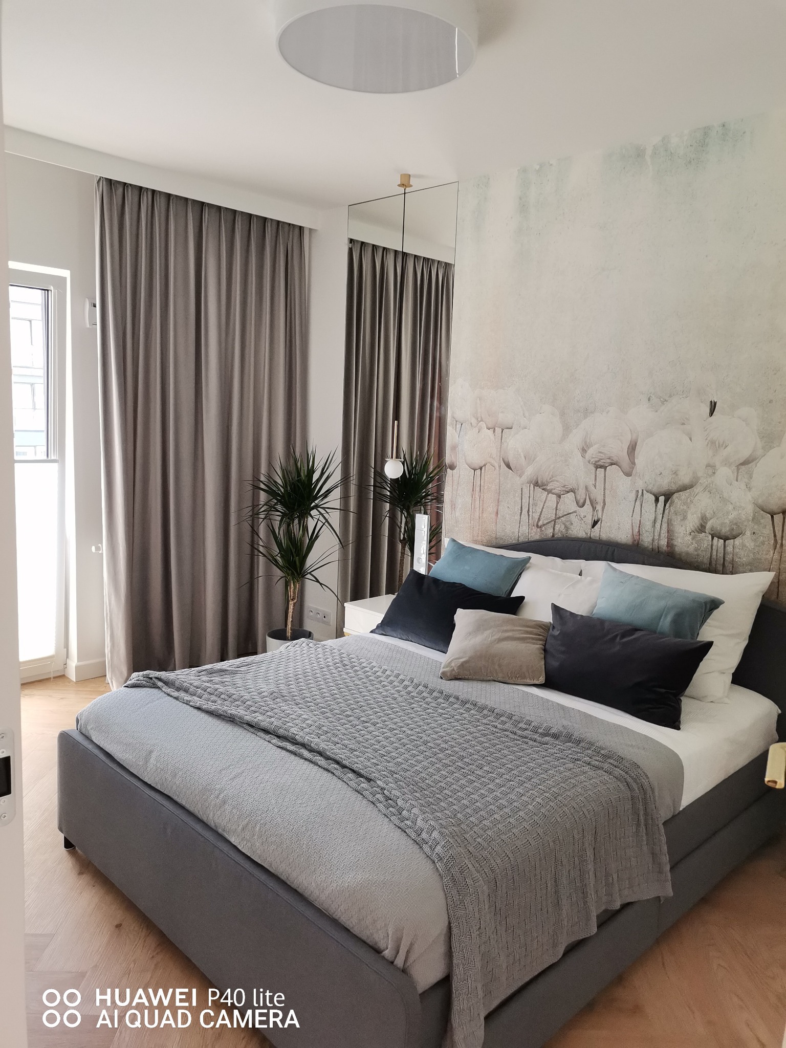 Jasna sypialnia z szarym łóżkiem, kocem o warkoczowym splocie i poduszkami w odcieniach szarości i błękitu, na ścianie fototapeta z motywem flamingów, obok okna szare zasłony, w rogu doniczka...