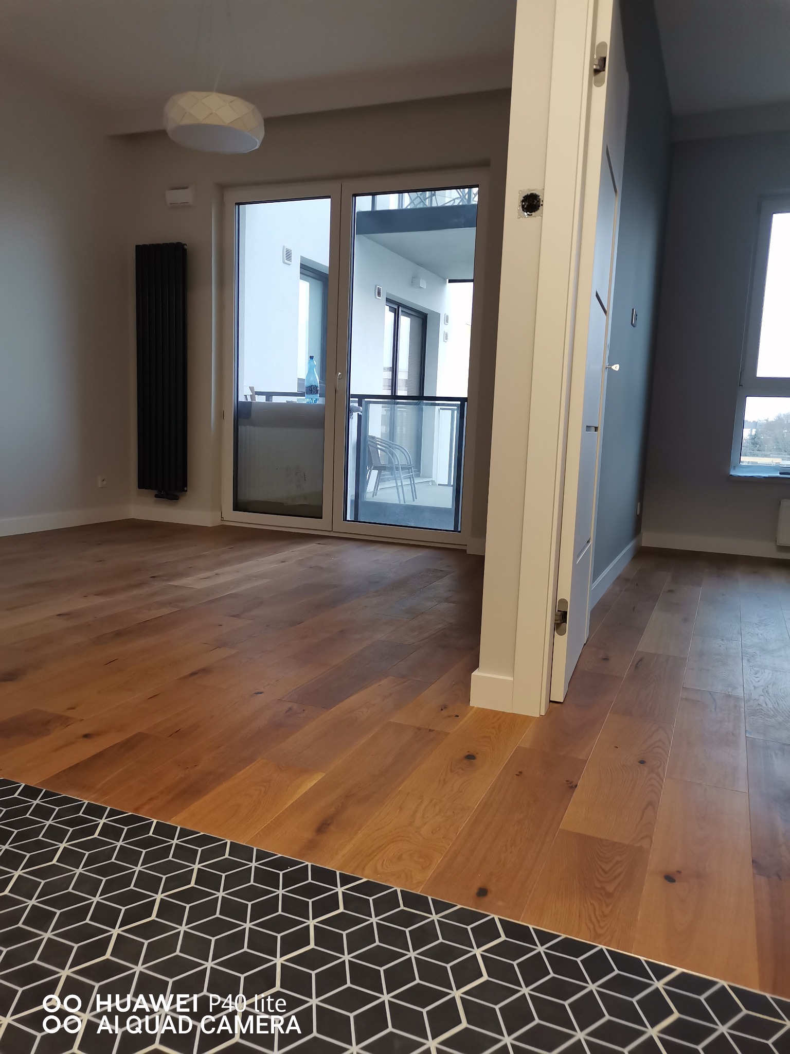 Nowoczesne wnętrze mieszkania z drewnianą podłogą, czarnym grzejnikiem panelowym i wyjściem na balkon. W tle widoczne okno i fragment ściany pomalowanej na szaro.