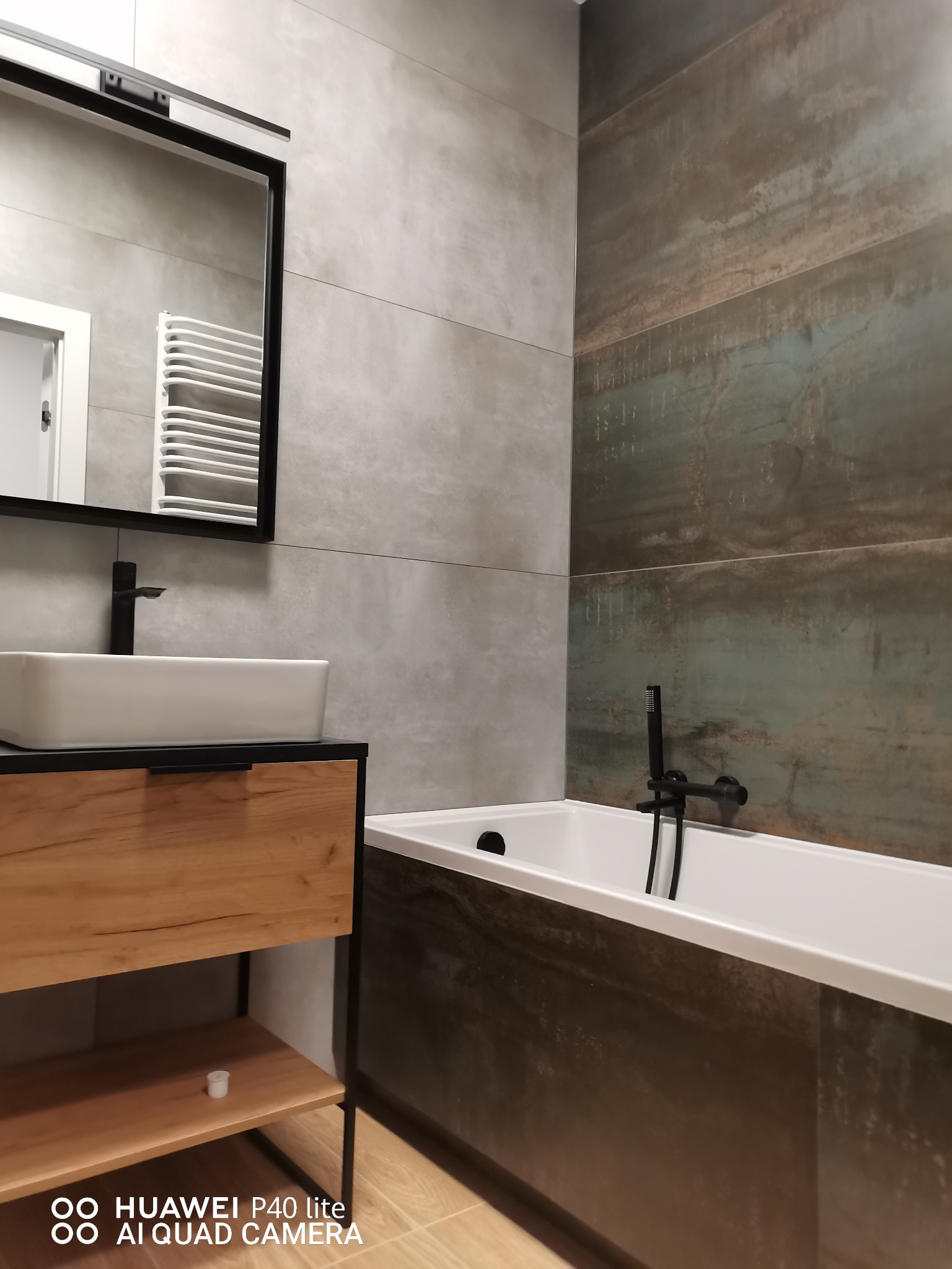 Nowoczesna łazienka z płytkami imitującymi beton i rdzę, czarna armatura, prostokątna umywalka nablatowa i grzejnik drabinkowy w lustrze.