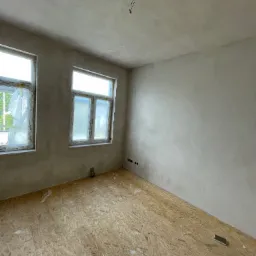 Pokój w trakcie remontu: ściany pokryte świeżą warstwą, widoczne niedoskonałości, podłoga z płyt OSB z resztkami zaprawy, zabezpieczone folią okna, wystające kable z sufitu.