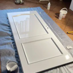 Białe drzwi z panelami, świeżo pomalowane na wysoki połysk, leżą na folii ochronnej. Obok puszka z farbą, wałek i pędzel. W tle drabina malarska.