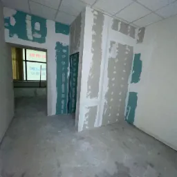 Wnętrze biura w trakcie remontu: ściany z płyt gipsowo-kartonowych z widocznymi miejscami łączeń i szpachlowania, podłoga betonowa z plamami, sufit podwieszany z kratką wentylacyjną.