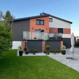 Dwukondygnacyjny dom jednorodzinny z elewacją w kolorach cegły, bieli i grafitu, z balkonem z szklaną balustradą, otoczony zadbanym trawnikiem, ozdobnymi krzewami w donicach i ścieżką z białego żwiru.