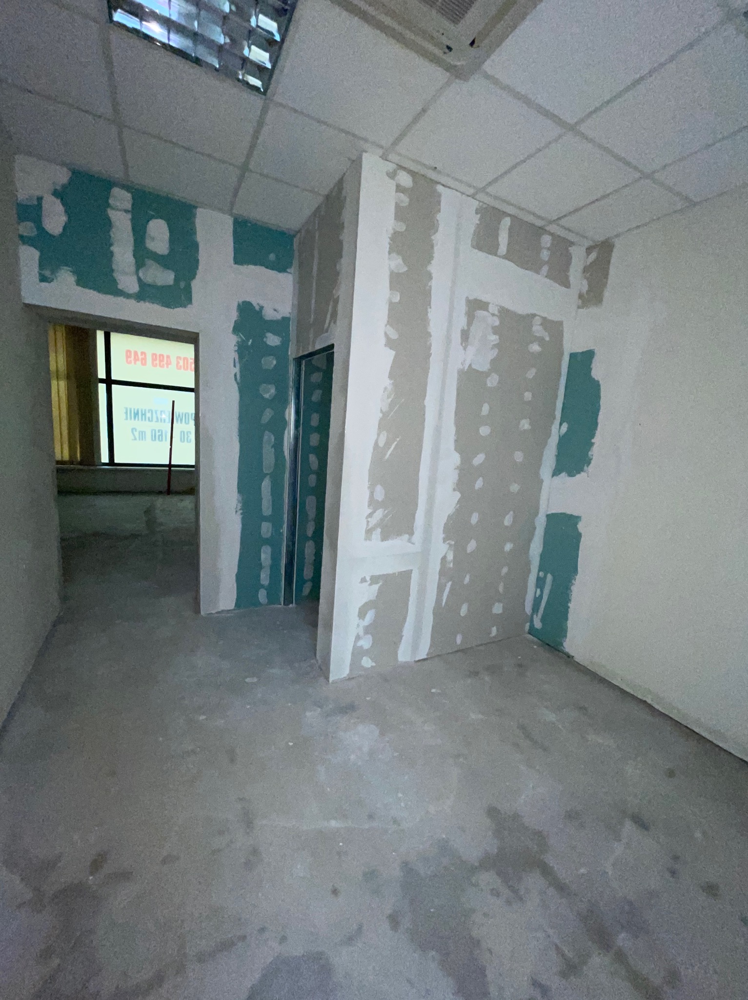 Wnętrze biura w trakcie remontu: ściany z płyt gipsowo-kartonowych z widocznymi miejscami łączeń i szpachlowania, podłoga betonowa z plamami, sufit podwieszany z kratką wentylacyjną.