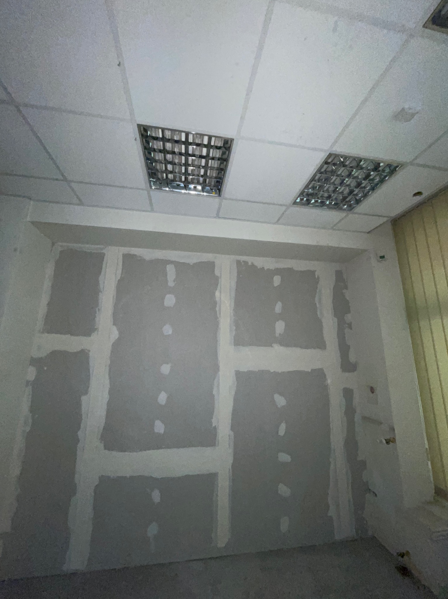 Ściana w trakcie przygotowania do malowania: płyty kartonowo-gipsowe z widocznymi łączeniami i zagipsowanymi wkrętami, sufit podwieszany z rastrowymi lampami.
