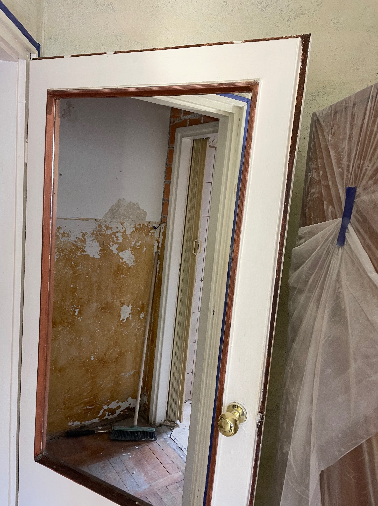 Przygotowanie do malowania futryny drzwiowej: świeżo pomalowana biała rama z brązowymi detalami, niebieska taśma malarska, ściana w tle z resztkami starej farby i widocznym fragmentem cegły...